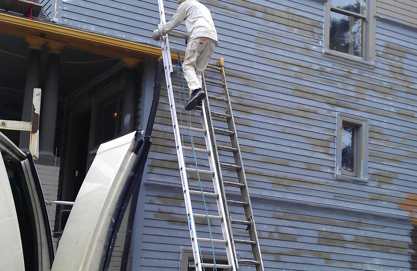 A painter on an extension ladder prepping tall siding on a Portland house
