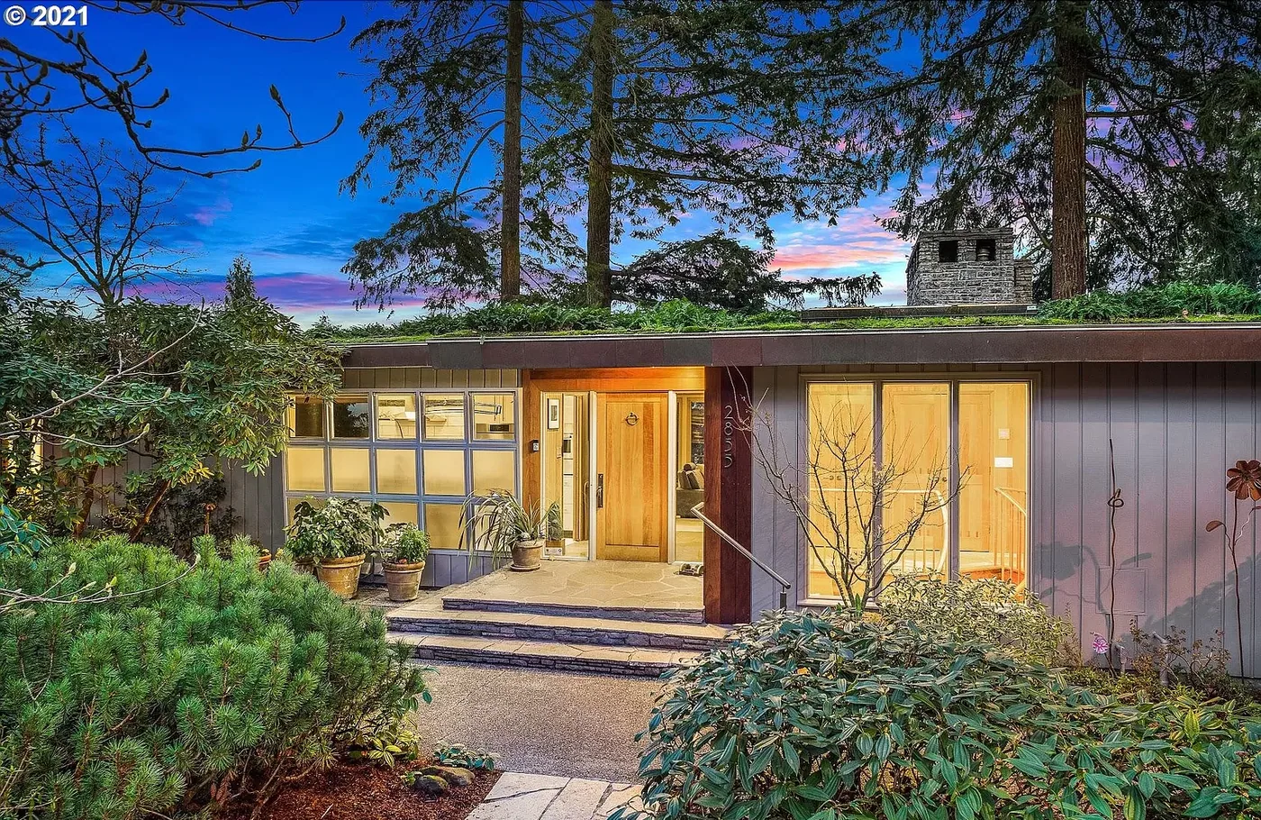 Mid-century home lit at dusk among tall trees